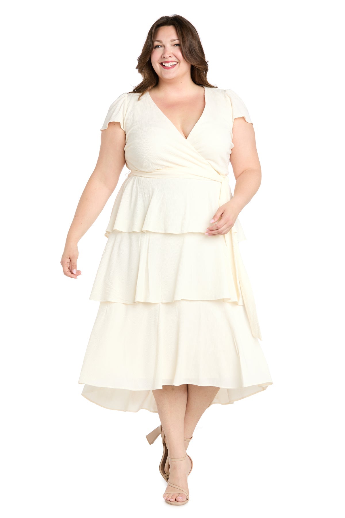 A woman in the R&M Richards 6677W Plus-Size White Party Dress with short sleeves and tiered ruffle layers stands against a white background, smiling in nude heels.